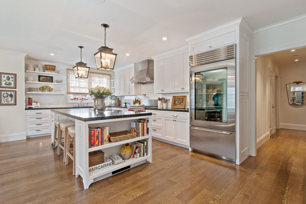 Inspiration for a traditional l-shaped kitchen in Santa Barbara with shaker cabinets, white cabinets, white splashback, stainless steel appliances, medium hardwood floors, with island, brown floor and black benchtop.