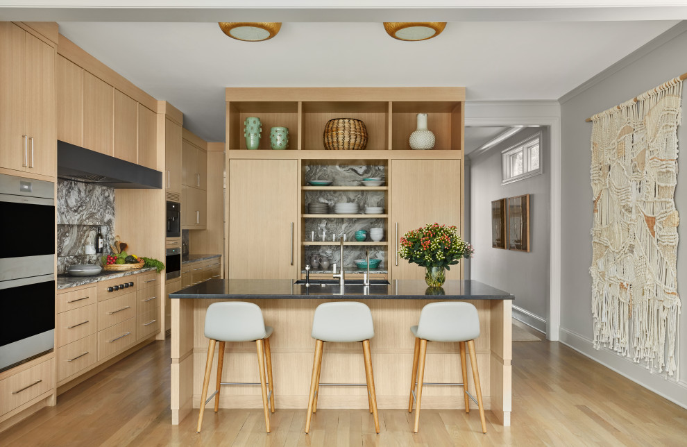 This is an example of a mid-sized contemporary l-shaped separate kitchen in Chicago with a drop-in sink, flat-panel cabinets, beige cabinets, marble benchtops, black splashback, marble splashback, panelled appliances, light hardwood floors, with island, beige floor and black benchtop.