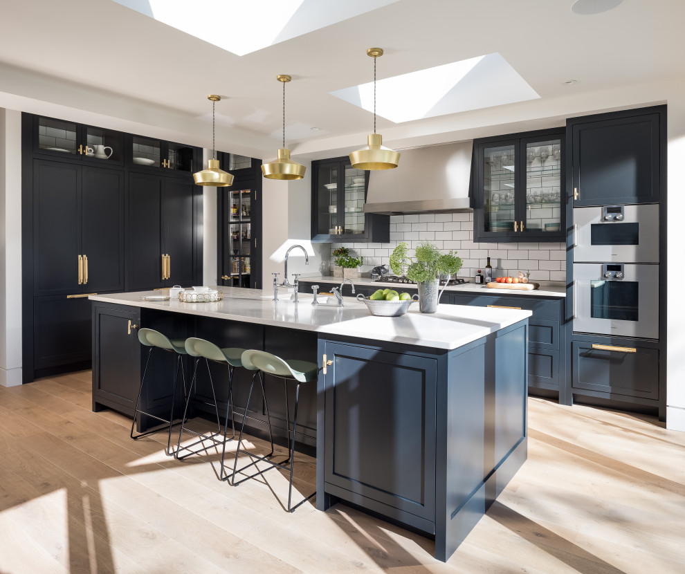Design ideas for a mid-sized transitional l-shaped kitchen in London with shaker cabinets, blue cabinets, granite benchtops, white splashback, subway tile splashback, light hardwood floors, with island, white benchtop, stainless steel appliances and beige floor.
