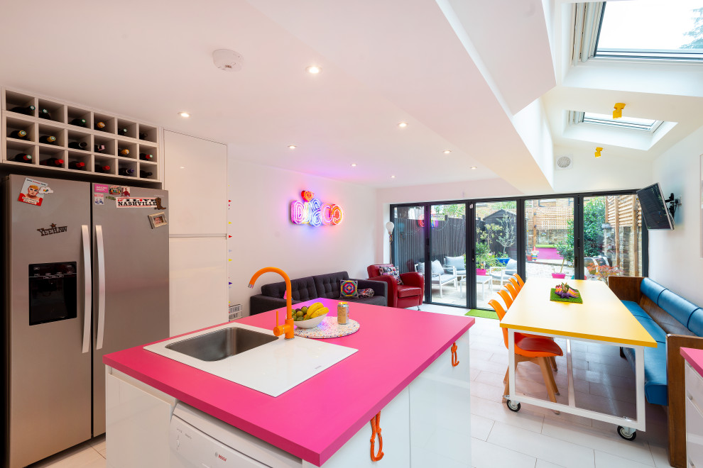 This is an example of a mid-sized eclectic l-shaped open plan kitchen in London with an integrated sink, flat-panel cabinets, white cabinets, solid surface benchtops, pink splashback, stainless steel appliances, ceramic floors, with island, white floor and pink benchtop.
