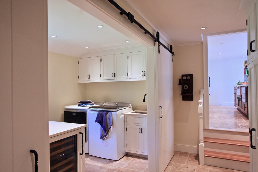 Mid-sized traditional single-wall utility room in New York with an undermount sink, white cabinets, travertine floors, a side-by-side washer and dryer, beige floor, shaker cabinets, solid surface benchtops and beige walls.