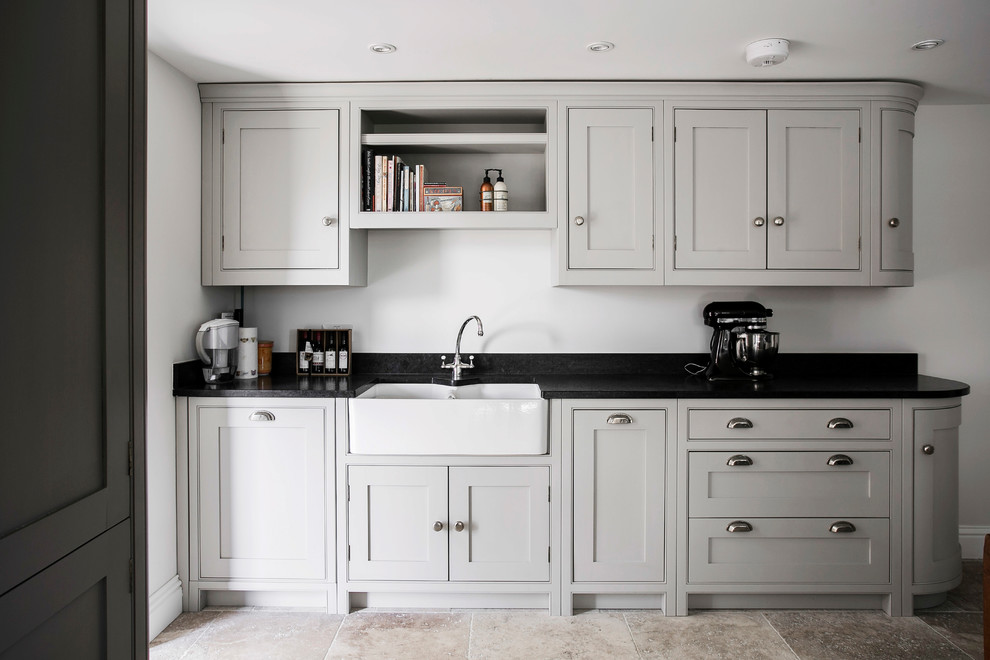 Mid-sized traditional u-shaped open plan kitchen in Kent with a farmhouse sink, shaker cabinets, grey cabinets, granite benchtops, brown splashback, ceramic splashback, black appliances, limestone floors, with island, beige floor and black benchtop.