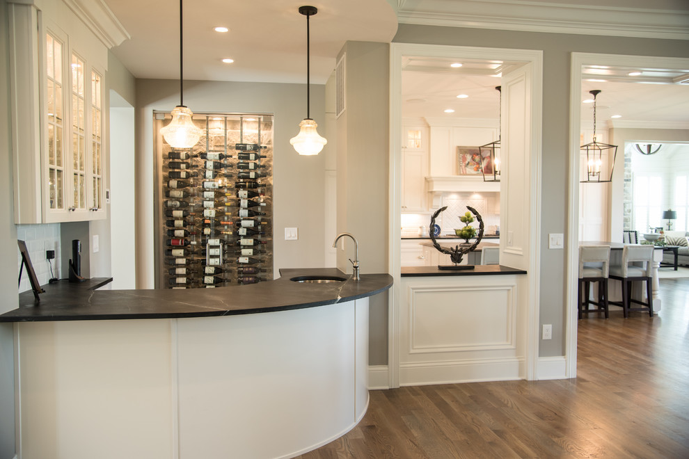 Design ideas for a mid-sized transitional galley wet bar in Cincinnati with an undermount sink, glass-front cabinets, white cabinets, soapstone benchtops, white splashback, subway tile splashback, medium hardwood floors and grey floor.