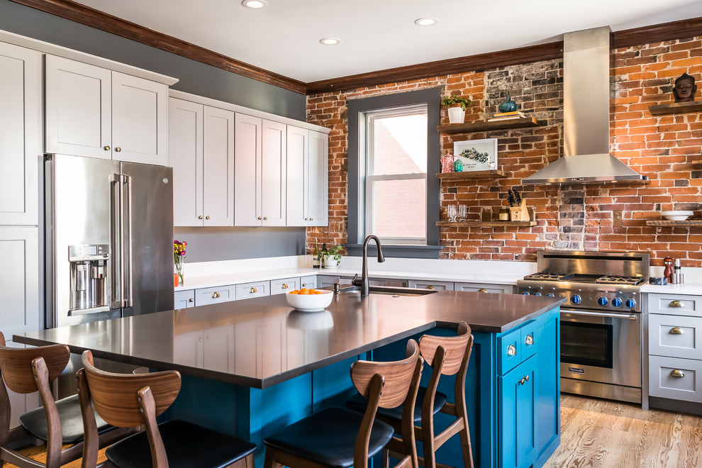 Mid-sized eclectic l-shaped kitchen in St Louis with a single-bowl sink, shaker cabinets, quartz benchtops, brick splashback, stainless steel appliances, medium hardwood floors, with island, white benchtop, blue cabinets and brown floor.