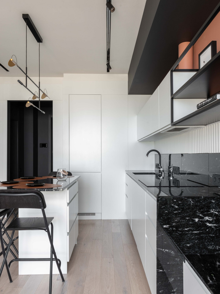 This is an example of a small scandinavian single-wall eat-in kitchen in Moscow with an undermount sink, flat-panel cabinets, white cabinets, granite benchtops, black splashback, granite splashback, black appliances, light hardwood floors, with island, grey floor and black benchtop.