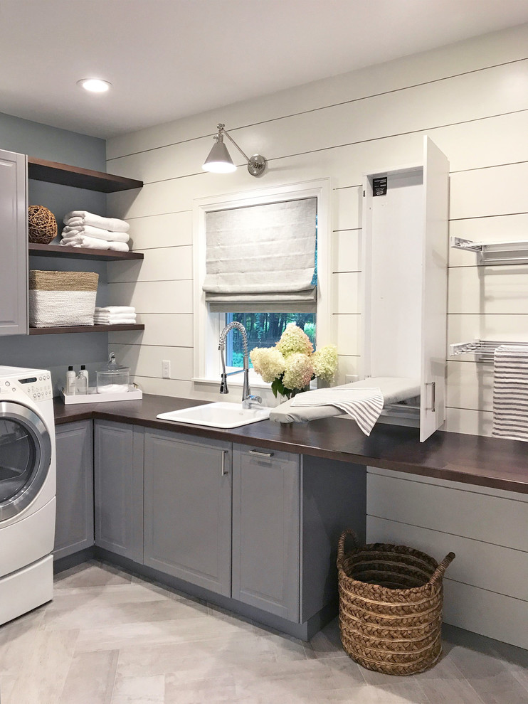 This is an example of a large traditional dedicated laundry room in New York with raised-panel cabinets, grey cabinets, wood benchtops, porcelain floors, a side-by-side washer and dryer, grey floor, brown benchtop, a drop-in sink and white walls.