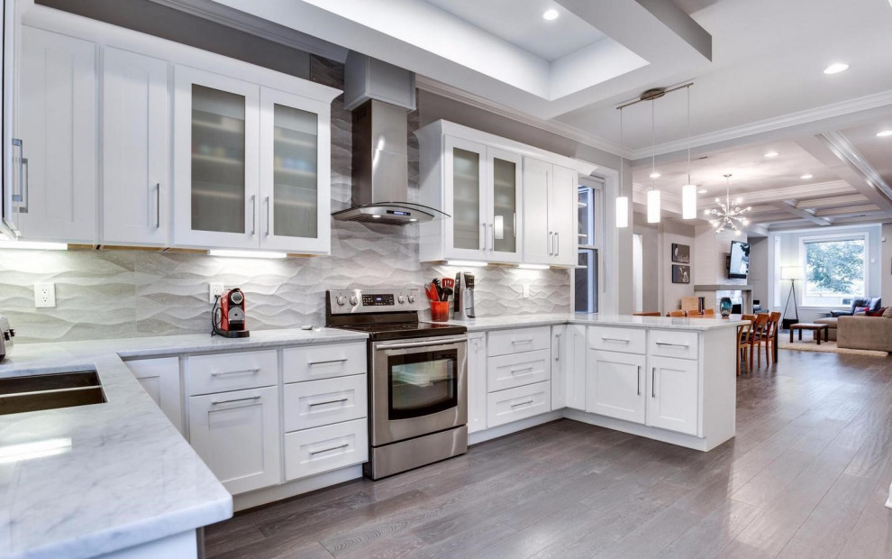 Modern inredning av ett stort vit vitt kök, med luckor med glaspanel, vita skåp, bänkskiva i kvarts, grått stänkskydd, rostfria vitvaror, mörkt trägolv, en köksö och brunt golv