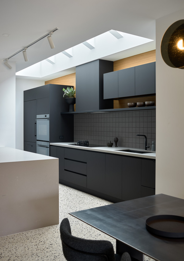 Large contemporary kitchen in Melbourne with flat-panel cabinets, black cabinets, solid surface benchtops, grey splashback, mosaic tile splashback, black appliances, concrete floors, with island, multi-coloured floor and grey benchtop.