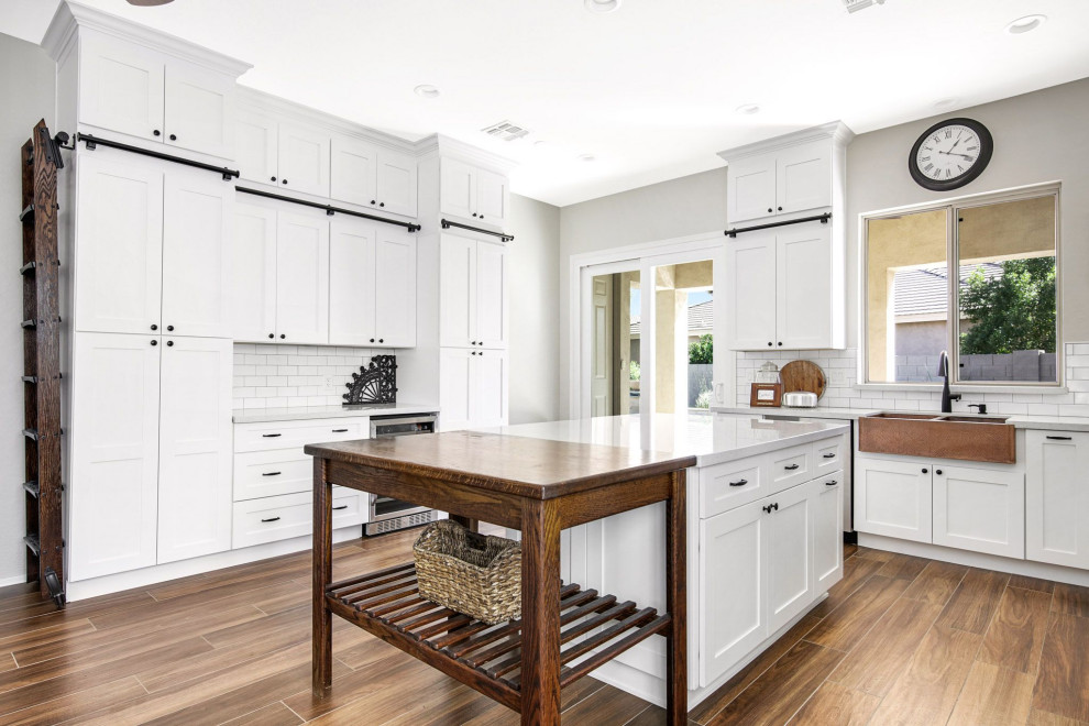 Photo of a mid-sized country kitchen in Phoenix with a farmhouse sink, shaker cabinets, white cabinets, white splashback, subway tile splashback, stainless steel appliances, with island and brown floor.