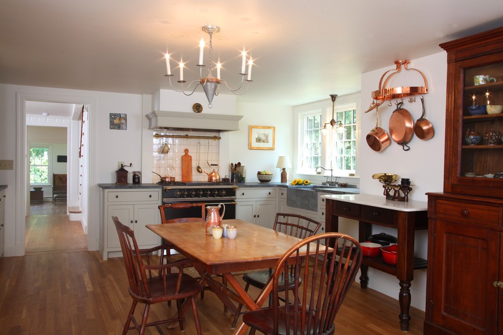 This is an example of a traditional l-shaped eat-in kitchen in Boston with a farmhouse sink, beaded inset cabinets, white cabinets, white splashback, soapstone benchtops, ceramic splashback, black appliances and medium hardwood floors.