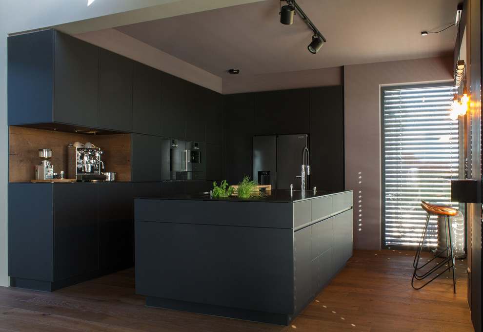 Design ideas for a large contemporary l-shaped open plan kitchen in Bremen with an undermount sink, flat-panel cabinets, black cabinets, solid surface benchtops, brown splashback, timber splashback, panelled appliances, medium hardwood floors, with island, brown floor and black benchtop.