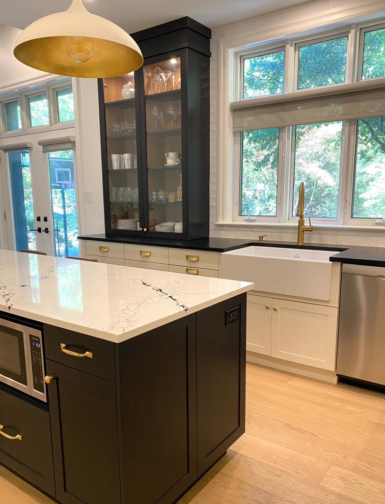 Mid-sized transitional l-shaped light wood floor open concept kitchen photo in Toronto with a farmhouse sink, shaker cabinets, white cabinets, quartz countertops, white backsplash, porcelain backsplash, an island and black countertops