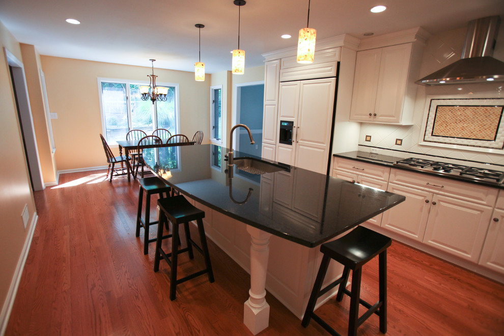 Mid-sized transitional l-shaped medium tone wood floor enclosed kitchen photo with an undermount sink, raised-panel cabinets, white cabinets, quartz countertops, white backsplash, ceramic backsplash, stainless steel appliances and an island