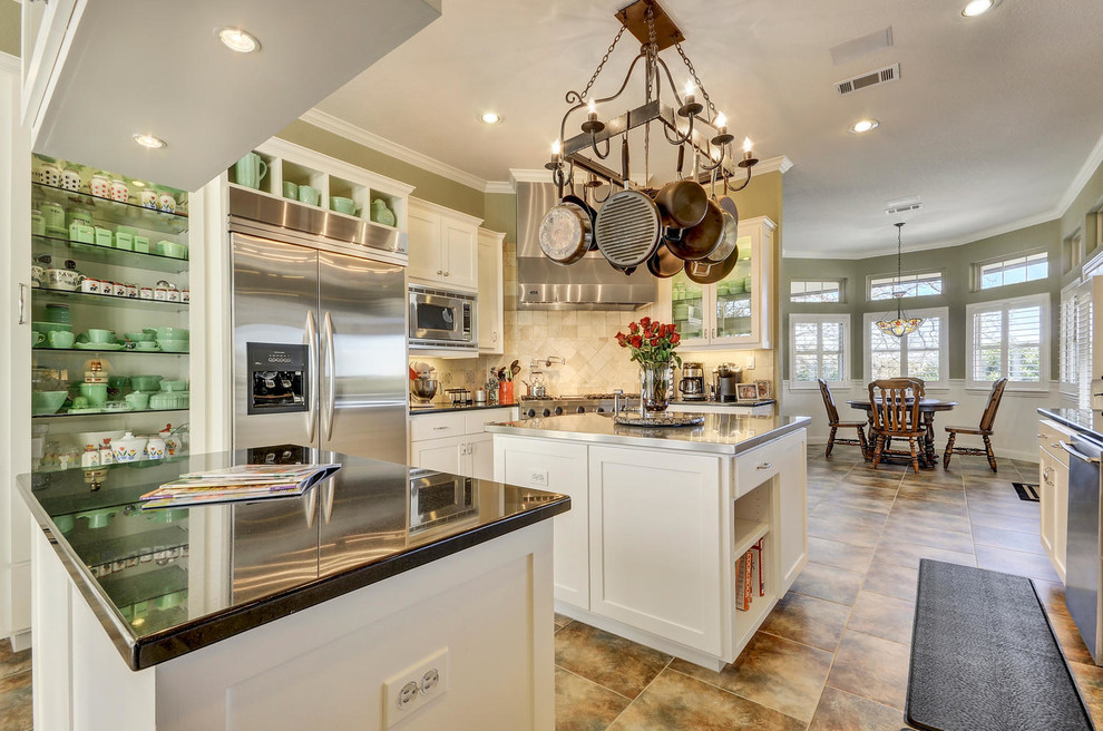 Photo of a mid-sized traditional kitchen in Austin with shaker cabinets, white cabinets, stainless steel benchtops, beige splashback, ceramic splashback, stainless steel appliances, ceramic floors, with island and brown floor.