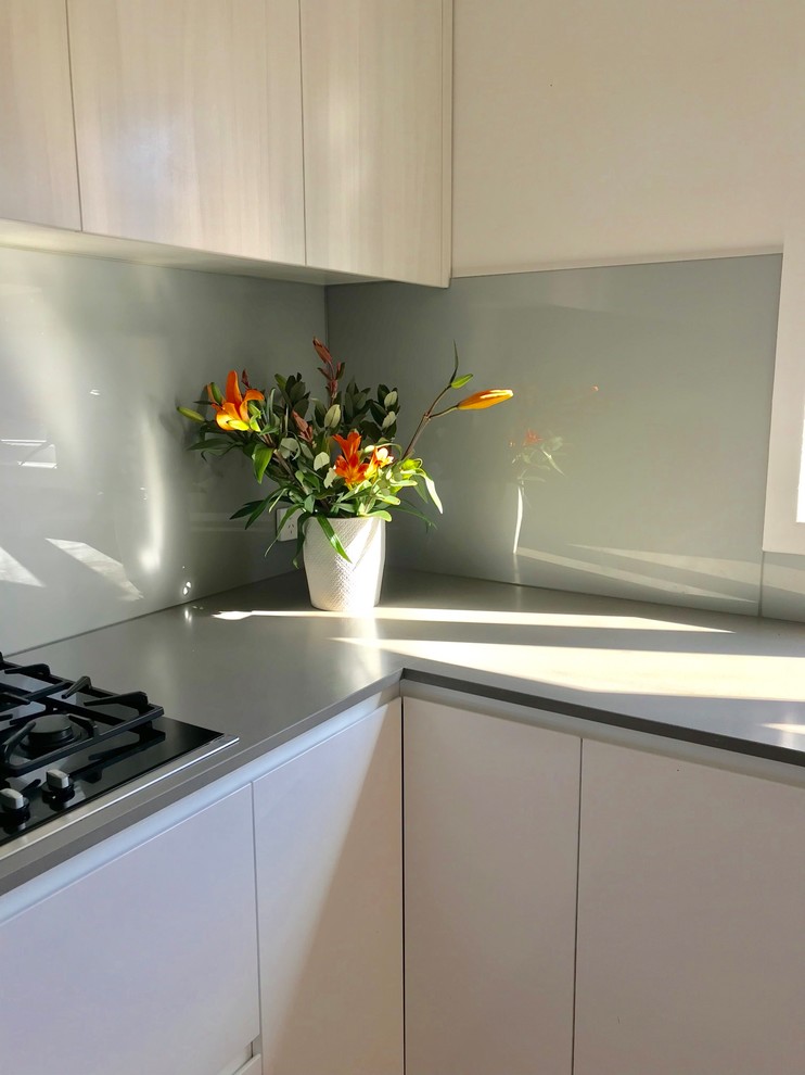 Mid-sized contemporary l-shaped eat-in kitchen in Melbourne with a double-bowl sink, white cabinets, marble benchtops, metallic splashback, glass sheet splashback, black appliances, light hardwood floors, with island, brown floor and grey benchtop.