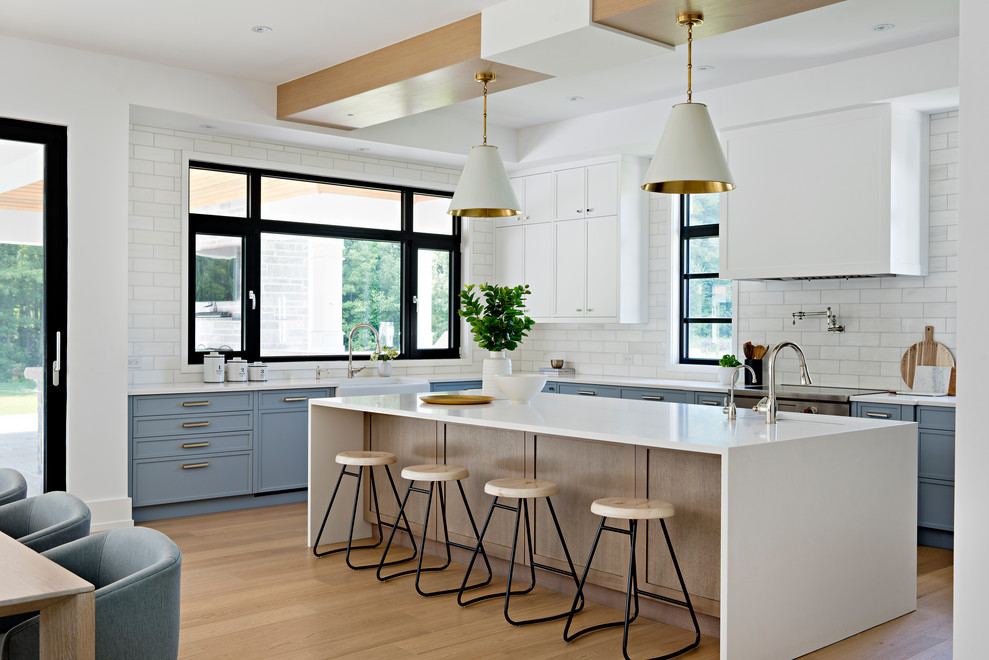 Transitional l-shaped eat-in kitchen in Toronto with a farmhouse sink, recessed-panel cabinets, blue cabinets, white splashback, subway tile splashback, stainless steel appliances, light hardwood floors, with island, beige floor and white benchtop.