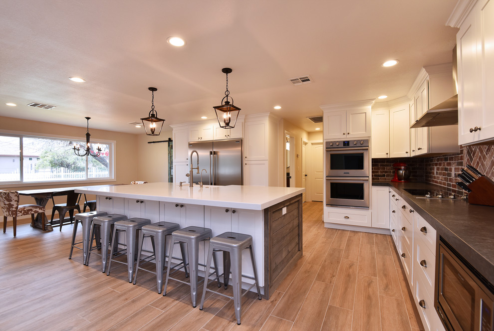 Design ideas for a large country l-shaped open plan kitchen in Phoenix with a farmhouse sink, shaker cabinets, white cabinets, quartz benchtops, red splashback, brick splashback, stainless steel appliances, light hardwood floors, with island and brown floor.