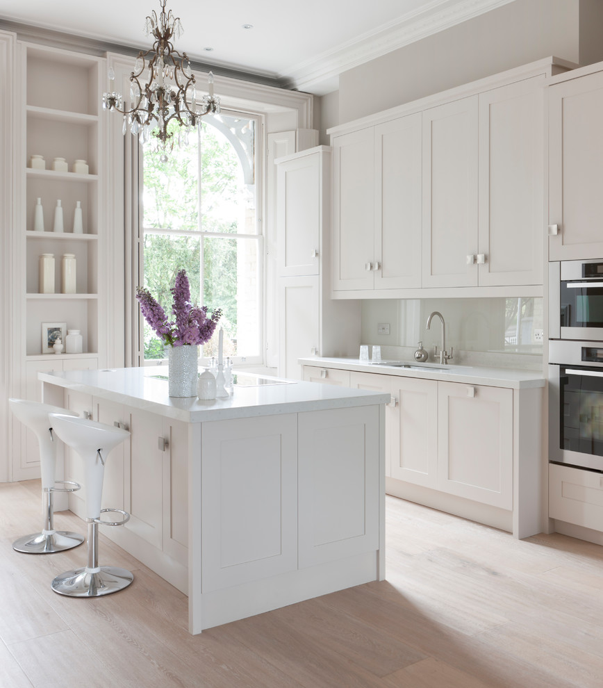 Design ideas for an expansive contemporary galley open plan kitchen in London with a drop-in sink, recessed-panel cabinets, beige cabinets, solid surface benchtops, white splashback, glass tile splashback, stainless steel appliances, light hardwood floors, multiple islands and white benchtop.