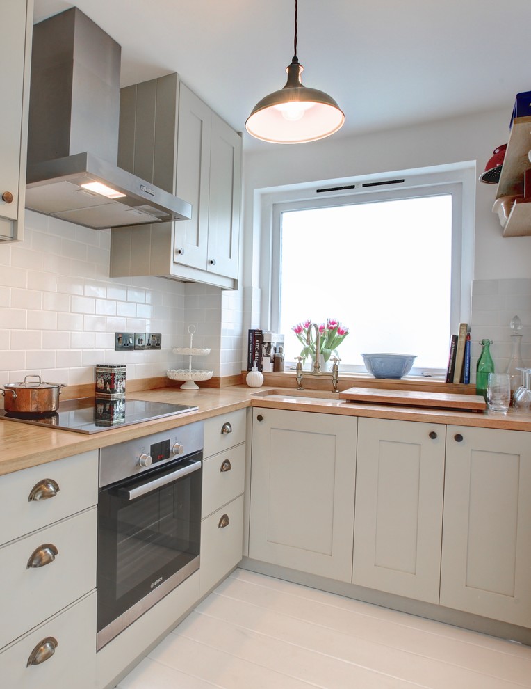 Photo of a small country l-shaped open plan kitchen in London with a drop-in sink, recessed-panel cabinets, grey cabinets, wood benchtops, white splashback, ceramic splashback, stainless steel appliances, painted wood floors and no island.