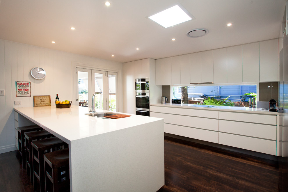 This is an example of a large contemporary galley eat-in kitchen in Brisbane with a double-bowl sink, flat-panel cabinets, white cabinets, quartz benchtops, brown splashback, glass tile splashback, stainless steel appliances, with island and dark hardwood floors.