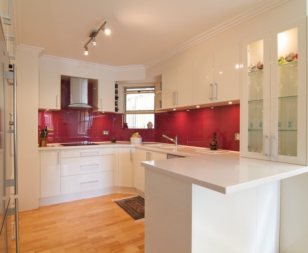 Bild på ett avskilt, litet funkis u-kök, med en integrerad diskho, släta luckor, vita skåp, bänkskiva i koppar, rött stänkskydd, glaspanel som stänkskydd, rostfria vitvaror och ljust trägolv