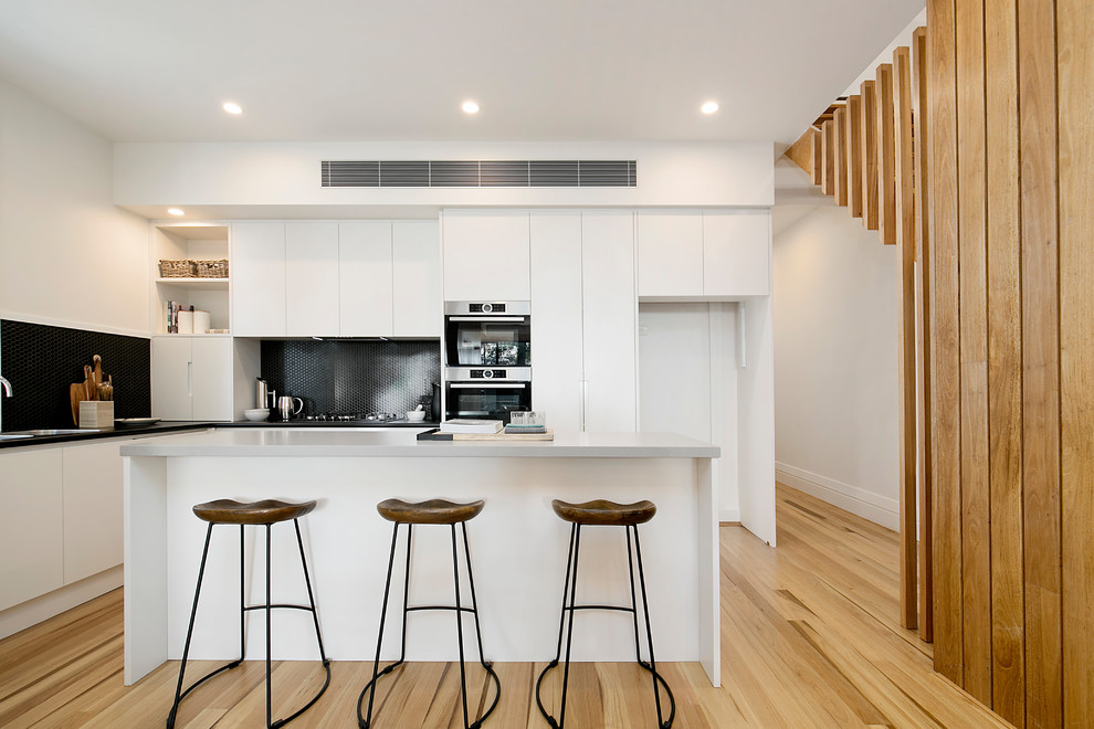 Photo of a contemporary l-shaped open plan kitchen in Sydney with black splashback, medium hardwood floors, with island, a drop-in sink, flat-panel cabinets, white cabinets, mosaic tile splashback and stainless steel appliances.