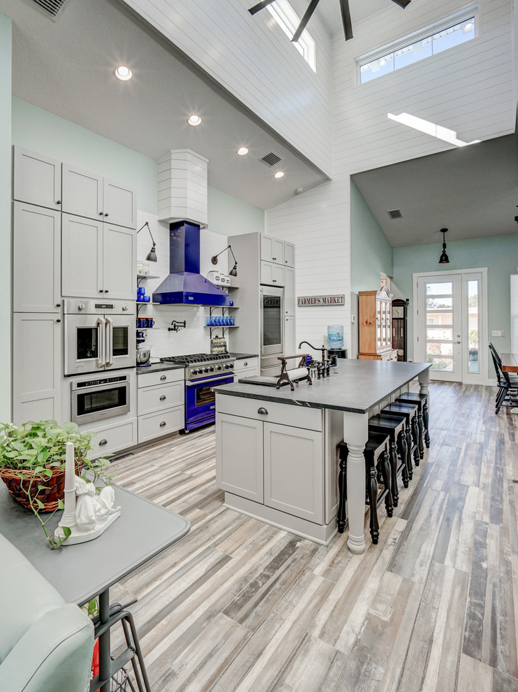 Photo of a mid-sized beach style galley open plan kitchen in Tampa with a farmhouse sink, recessed-panel cabinets, grey cabinets, quartzite benchtops, white splashback, ceramic splashback, stainless steel appliances, ceramic floors, with island, grey floor and grey benchtop.