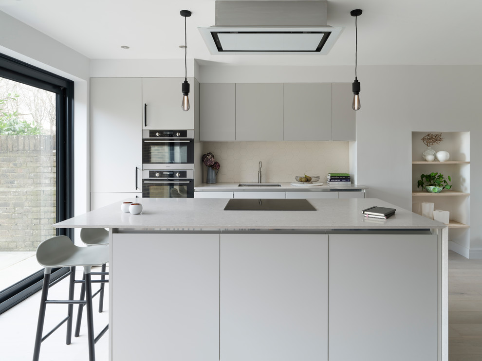 This is an example of a mid-sized contemporary single-wall open plan kitchen in Sussex with a drop-in sink, flat-panel cabinets, grey cabinets, beige splashback, ceramic splashback, light hardwood floors, with island and white floor.