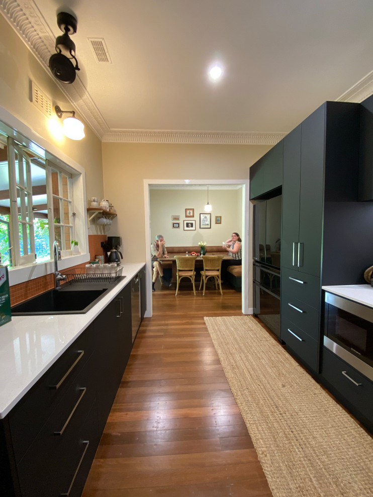This is an example of a small contemporary galley eat-in kitchen in Brisbane with a double-bowl sink, flat-panel cabinets, black cabinets, quartz benchtops, orange splashback, terra-cotta splashback, black appliances, medium hardwood floors, brown floor and white benchtop.