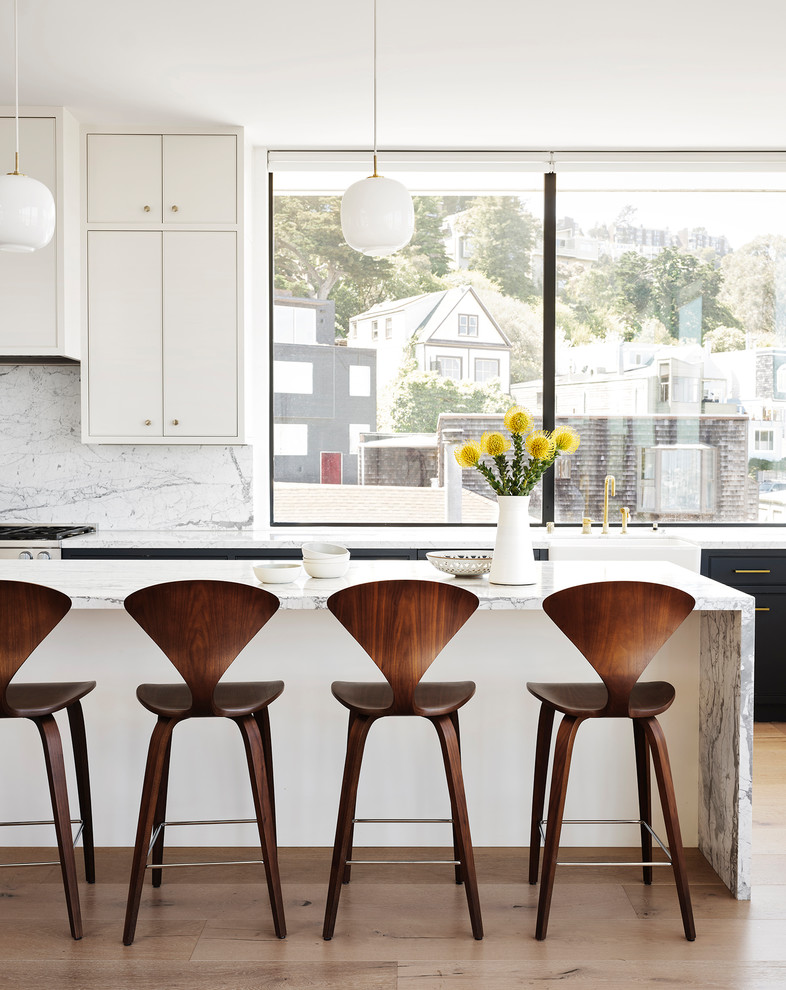 This is an example of a large midcentury open plan kitchen in San Francisco with a farmhouse sink, flat-panel cabinets, white cabinets, white splashback, stone slab splashback, light hardwood floors, with island, beige floor, white benchtop and marble benchtops.