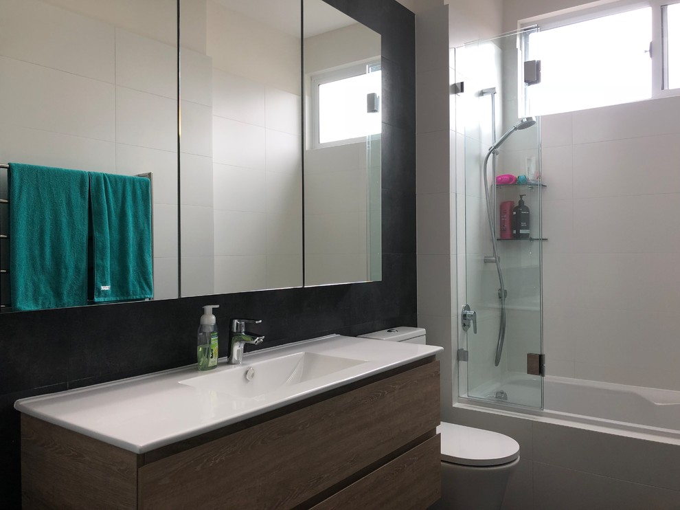 Photo of a small modern master bathroom in Sydney with flat-panel cabinets, medium wood cabinets, a drop-in tub, a shower/bathtub combo, a one-piece toilet, black and white tile, porcelain tile, white walls, a console sink, solid surface benchtops, black floor and white benchtops.