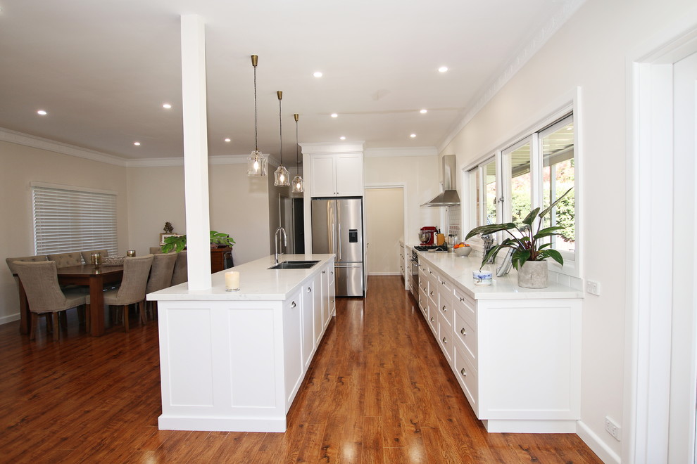 Large transitional galley open plan kitchen in Sydney with a double-bowl sink, shaker cabinets, white cabinets, quartz benchtops, white splashback, subway tile splashback, stainless steel appliances, medium hardwood floors, with island, brown floor and white benchtop.