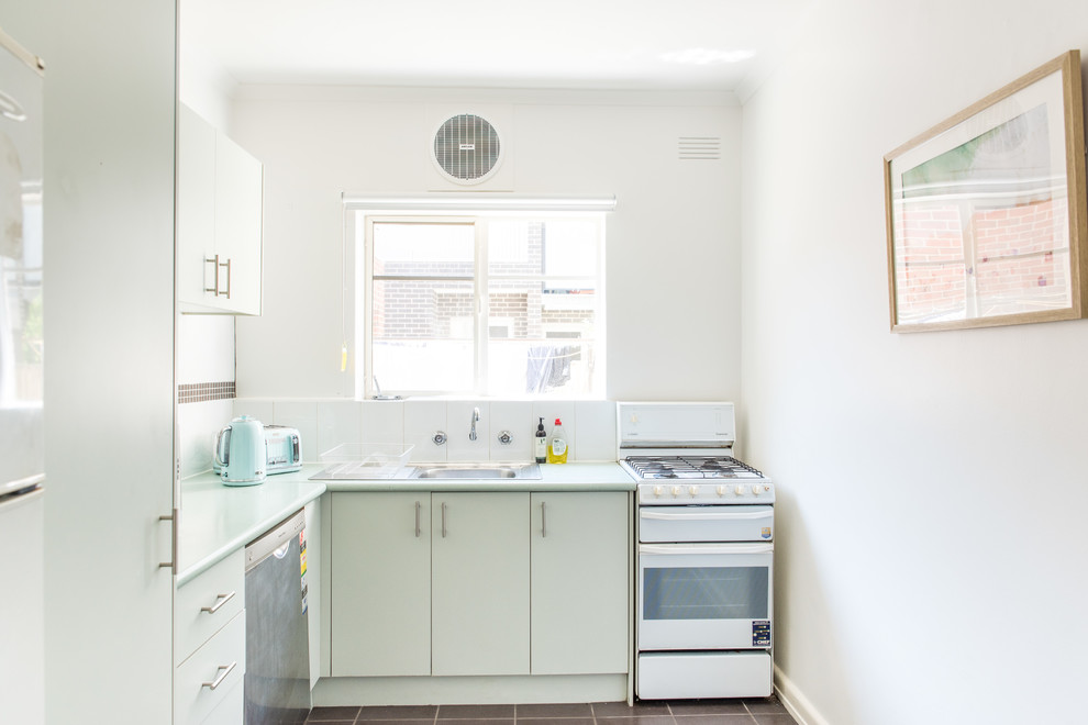 This is an example of a beach style l-shaped separate kitchen in Melbourne with a drop-in sink, flat-panel cabinets, white cabinets, white splashback, stainless steel appliances, no island, brown floor and white benchtop.