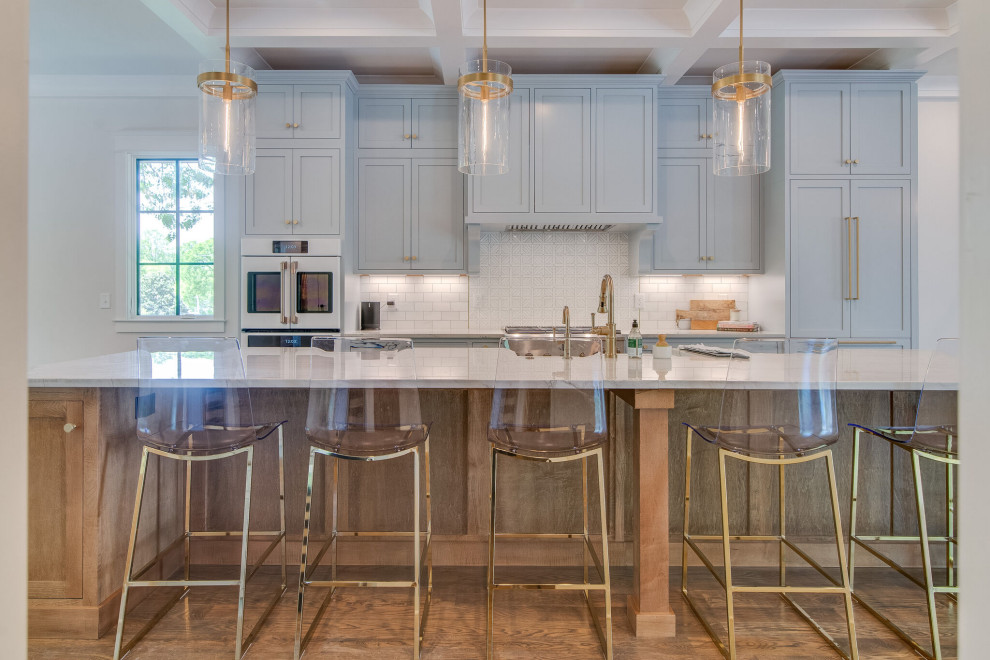 Large transitional galley open plan kitchen in Nashville with a farmhouse sink, shaker cabinets, grey cabinets, quartzite benchtops, white splashback, stone tile splashback, white appliances, medium hardwood floors, with island, brown floor, grey benchtop and coffered.