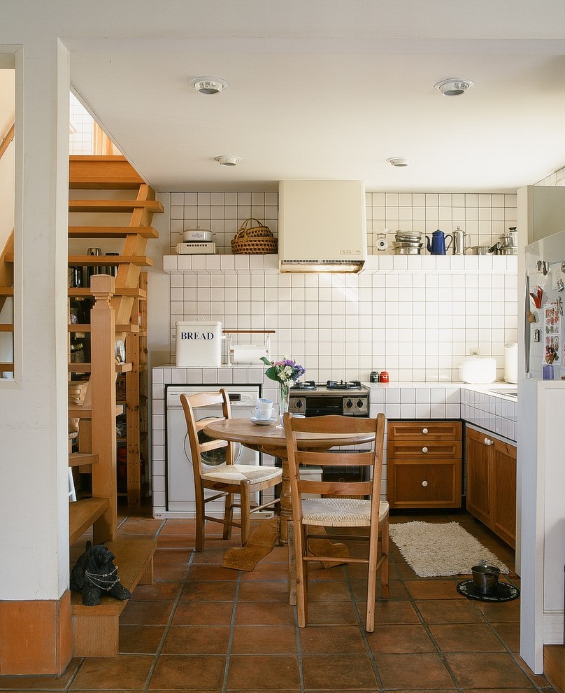 This is an example of a small country l-shaped eat-in kitchen in Other with raised-panel cabinets, medium wood cabinets, no island, tile benchtops, white appliances and brick floors.