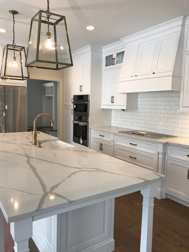 Inspiration for a mid-sized country kitchen in Cleveland with a farmhouse sink, recessed-panel cabinets, white cabinets, quartzite benchtops, white splashback, subway tile splashback, stainless steel appliances, medium hardwood floors, with island and brown floor.