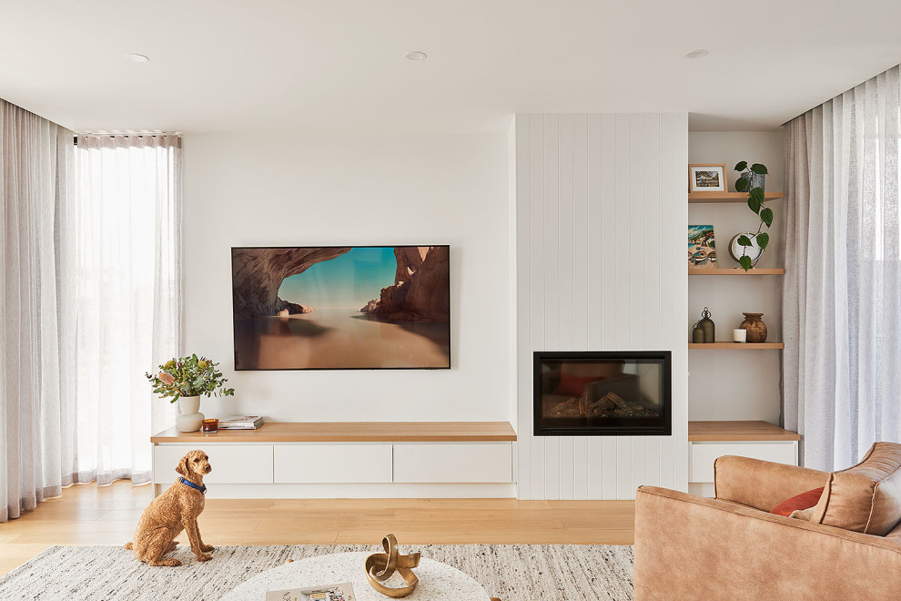 Photo of a large beach style open concept family room in Geelong with white walls, light hardwood floors, a standard fireplace, a wood fireplace surround, a wall-mounted tv and brown floor.