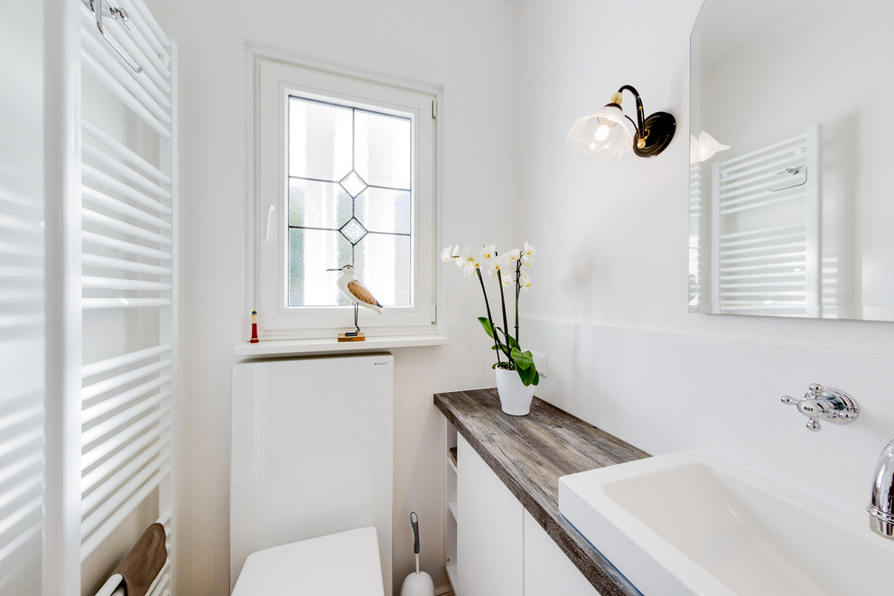 Photo of a small beach style powder room in Hamburg with flat-panel cabinets, white cabinets, a wall-mount toilet, white tile, mirror tile, white walls, ceramic floors, a vessel sink, beige floor, wood benchtops and brown benchtops.
