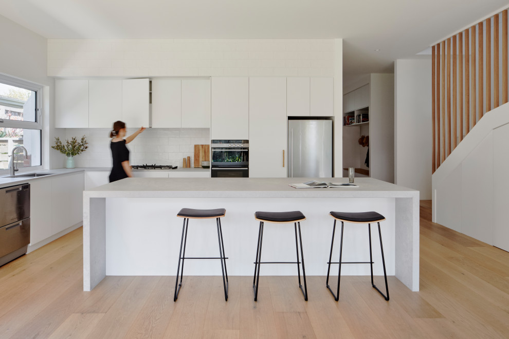 Photo of a scandinavian open plan kitchen in Melbourne with a single-bowl sink, flat-panel cabinets, white cabinets, solid surface benchtops, white splashback, ceramic splashback, stainless steel appliances, light hardwood floors, with island and white benchtop.