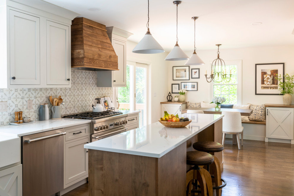Photo of a large farmhouse l-shaped kitchen/diner in New York with a belfast sink, shaker cabinets, grey cabinets, engineered stone countertops, beige splashback, ceramic splashback, stainless steel appliances, medium hardwood flooring, multiple islands, brown floors and white worktops.