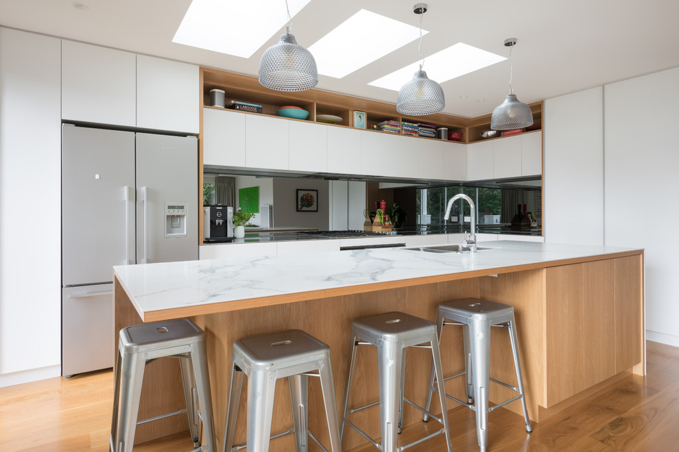 Inspiration for a mid-sized contemporary l-shaped eat-in kitchen in Auckland with a double-bowl sink, recessed-panel cabinets, white cabinets, granite benchtops, metallic splashback, mirror splashback, stainless steel appliances, medium hardwood floors, with island, brown floor and white benchtop.