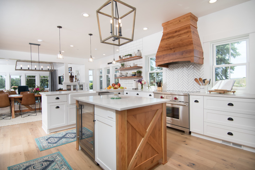 This is an example of a large country l-shaped eat-in kitchen in Portland with a farmhouse sink, white cabinets, white splashback, stainless steel appliances, with island, shaker cabinets, light hardwood floors, beige floor and grey benchtop.