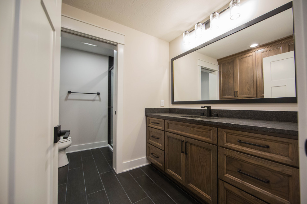 Large classic shower room bathroom in Indianapolis with recessed-panel cabinets, brown cabinets, a two-piece toilet, beige walls, porcelain flooring, a submerged sink, granite worktops, black floors, a hinged door, black worktops, a single sink and a freestanding vanity unit.