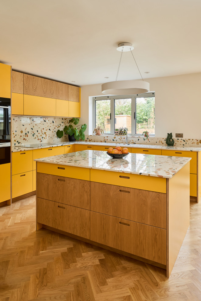 This is an example of a large retro l-shaped kitchen/diner in Kent with a submerged sink, flat-panel cabinets, yellow cabinets, terrazzo worktops, multi-coloured splashback, stone slab splashback, stainless steel appliances, medium hardwood flooring, an island, brown floors and multicoloured worktops.