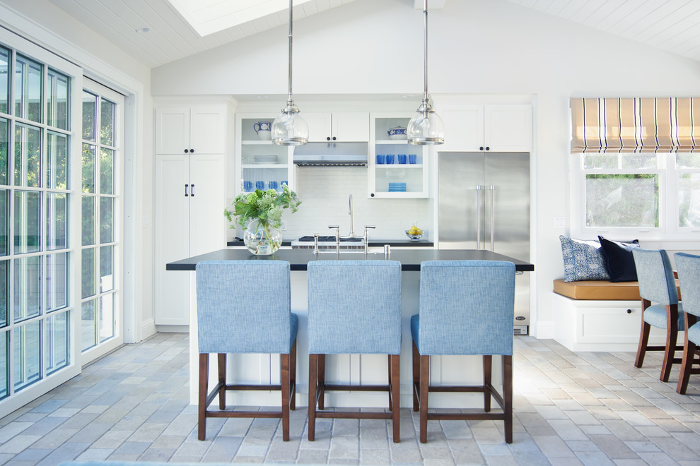 This is an example of a beach style eat-in kitchen in San Francisco with glass-front cabinets, white cabinets, white splashback, stainless steel appliances, with island and beige floor.
