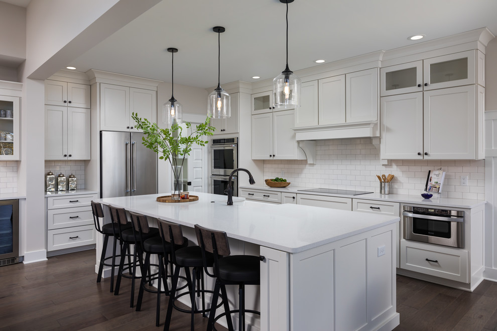 Mid-sized beach style galley eat-in kitchen in Grand Rapids with a farmhouse sink, recessed-panel cabinets, white cabinets, quartzite benchtops, white splashback, subway tile splashback, stainless steel appliances, medium hardwood floors, with island, brown floor and white benchtop.