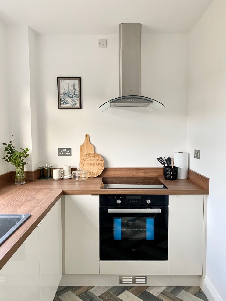 Small contemporary l-shaped separate kitchen in Gloucestershire with an integrated sink, flat-panel cabinets, white cabinets, wood benchtops, brown splashback, timber splashback, black appliances, linoleum floors, no island, multi-coloured floor and brown benchtop.