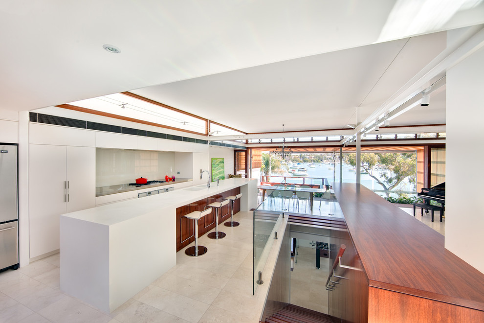 Photo of an expansive modern single-wall open plan kitchen in Sydney with an integrated sink, flat-panel cabinets, white cabinets, stainless steel benchtops, white splashback, glass sheet splashback, stainless steel appliances, travertine floors and with island.