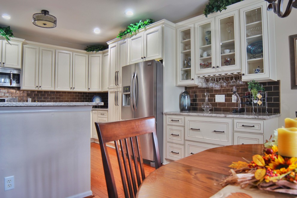 Example of a mid-sized u-shaped medium tone wood floor and brown floor kitchen design in Charleston with an undermount sink, raised-panel cabinets, beige cabinets, granite countertops, brown backsplash, ceramic backsplash, stainless steel appliances and beige countertops