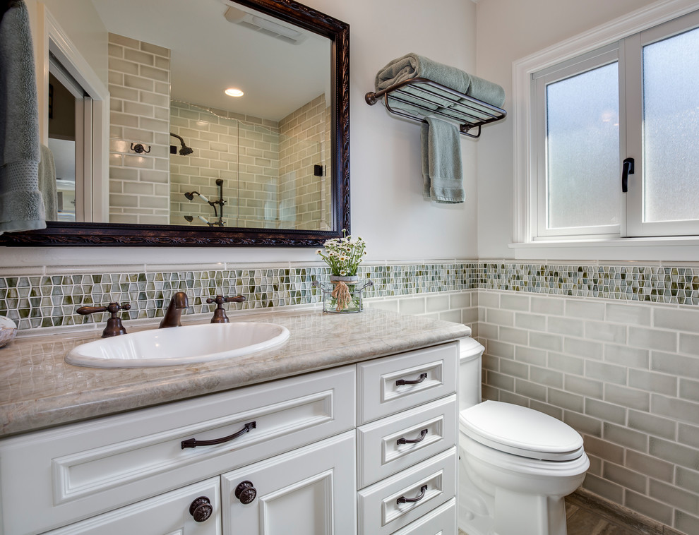 Design ideas for a mid-sized traditional 3/4 bathroom in Los Angeles with recessed-panel cabinets, white cabinets, gray tile, subway tile, white walls, a drop-in sink, an alcove shower, a two-piece toilet, engineered quartz benchtops, a hinged shower door and beige benchtops.